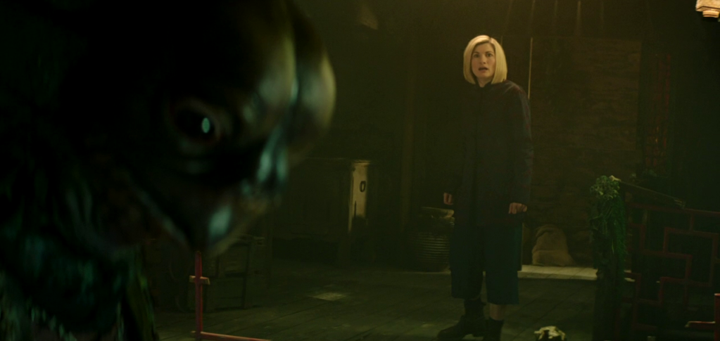 Foreground left: the head of a Sea Devil, a human-sized amphibian creature, staring down at controls (out fo shot); background centre right, a blonde-haired, pale-skinned woman - Jodie Whittaker playing the Doctor - look on in horror and disgust.